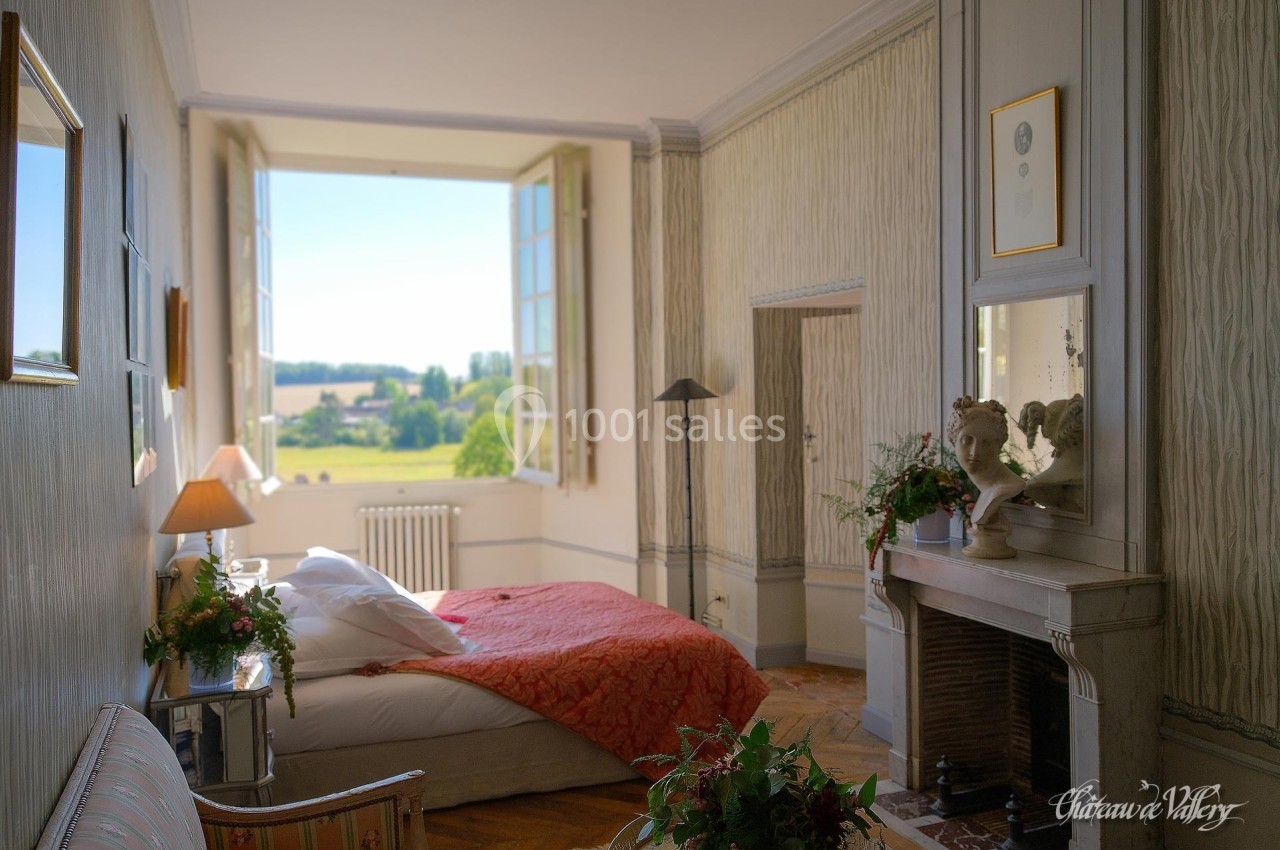 Chambre lumineuse avec lit double, cheminée, décor classique et vue sur un paysage verdoyant par une grande fenêtre ouverte.