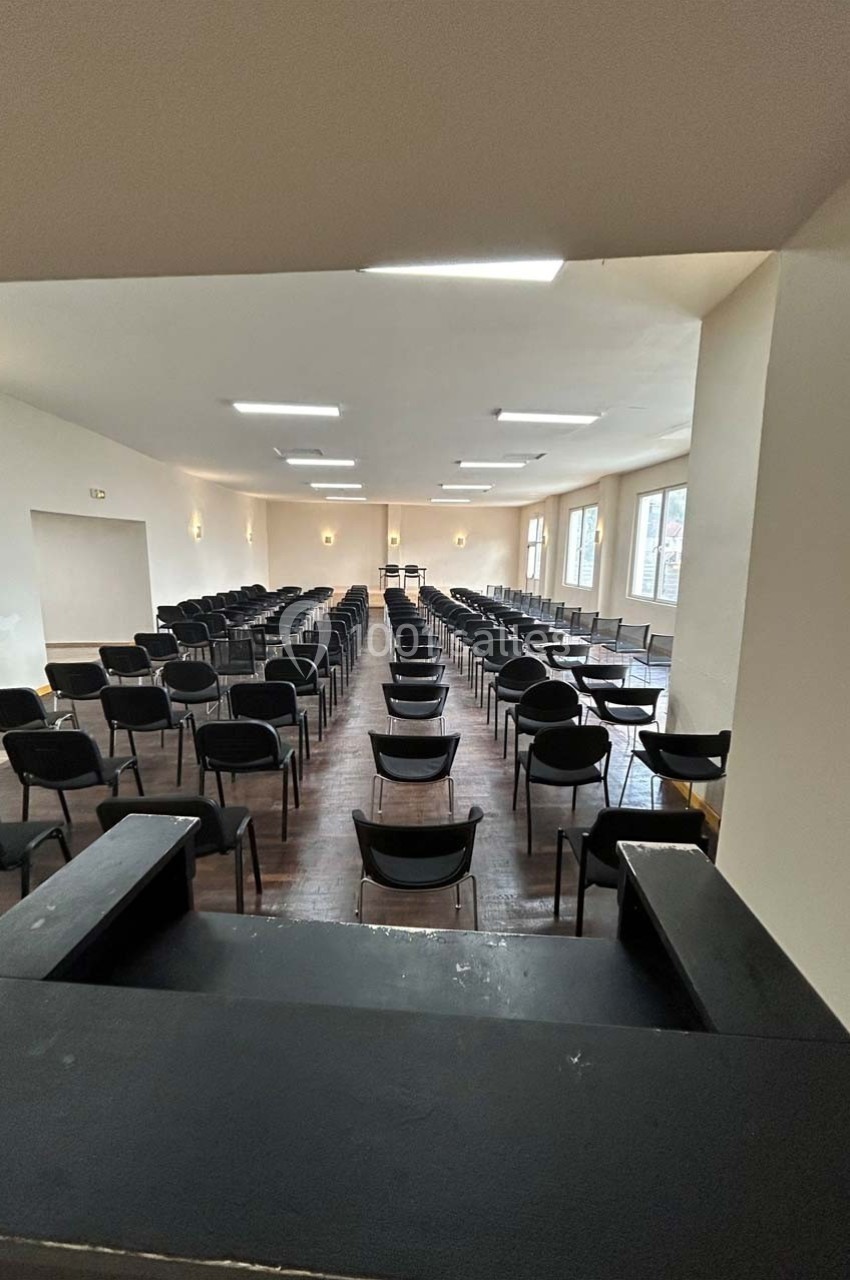 Salle vide avec des rangées de chaises noires alignées, éclairée par des fenêtres et des plafonniers.