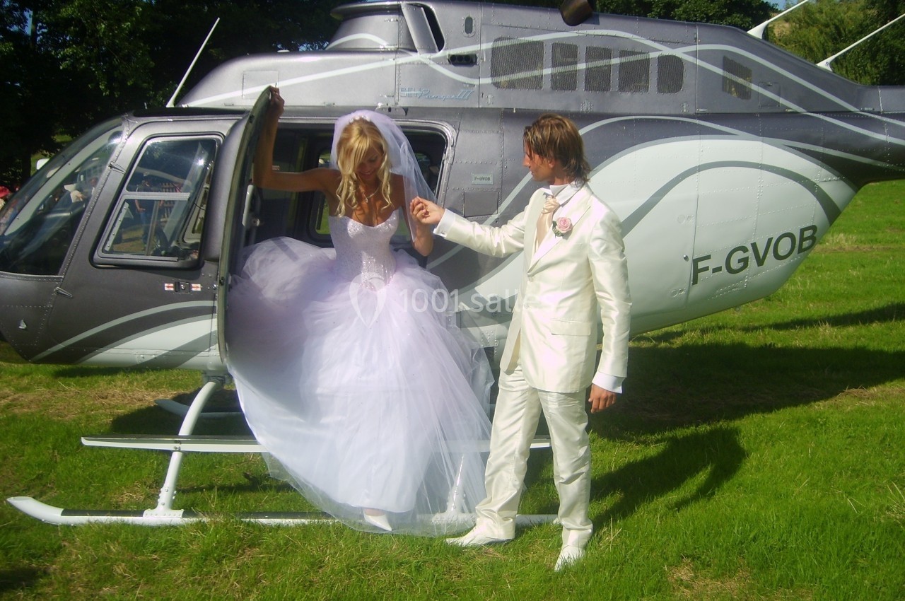 Une mariée en robe blanche descend d'un hélicoptère, aidée par un homme en costume clair, sur une pelouse verte.