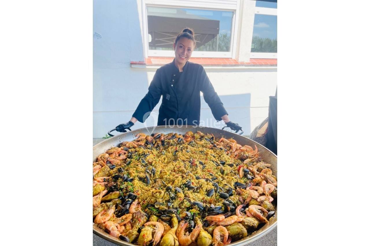 Une cheffe souriante présente une grande paella garnie de fruits de mer, poulet et légumes, en extérieur.
