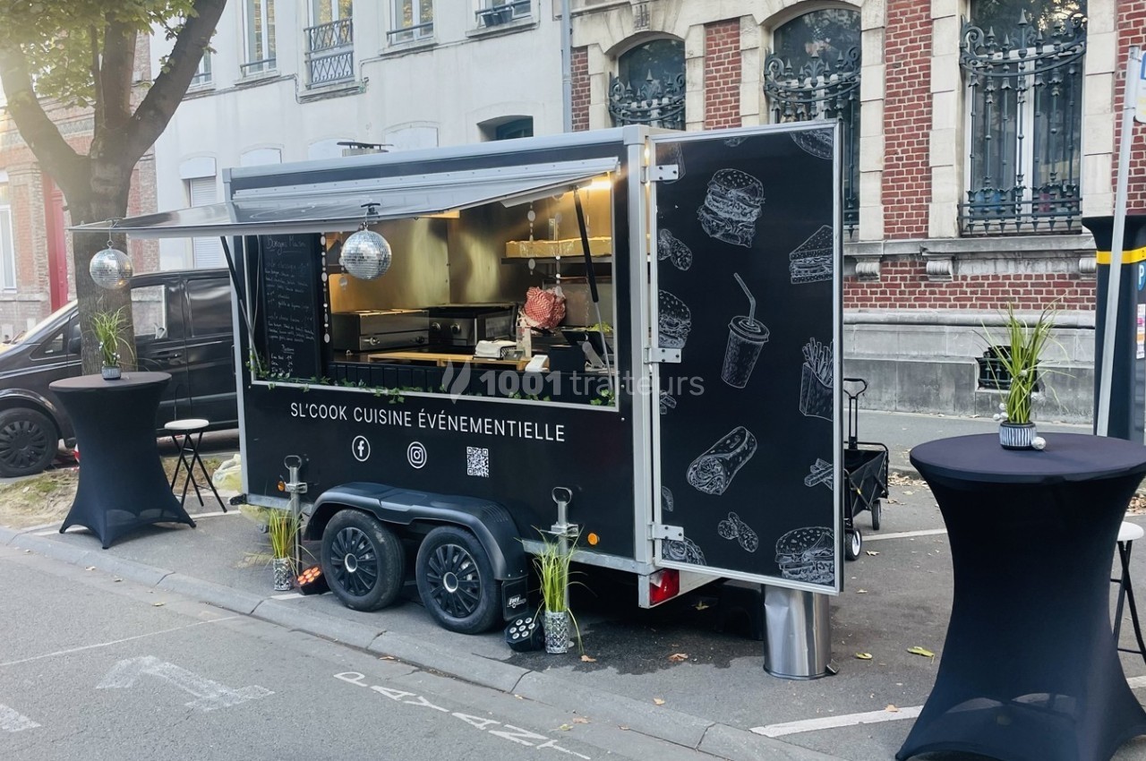 Food truck noir stationné dans une rue, avec illustrations de plats sur sa façade et tables hautes à proximité.