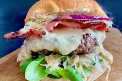 Burger au saumon fumé avec laitue, tomate et sauce, posé sur une table en marbre avec citron vert et herbes en arrière-plan.