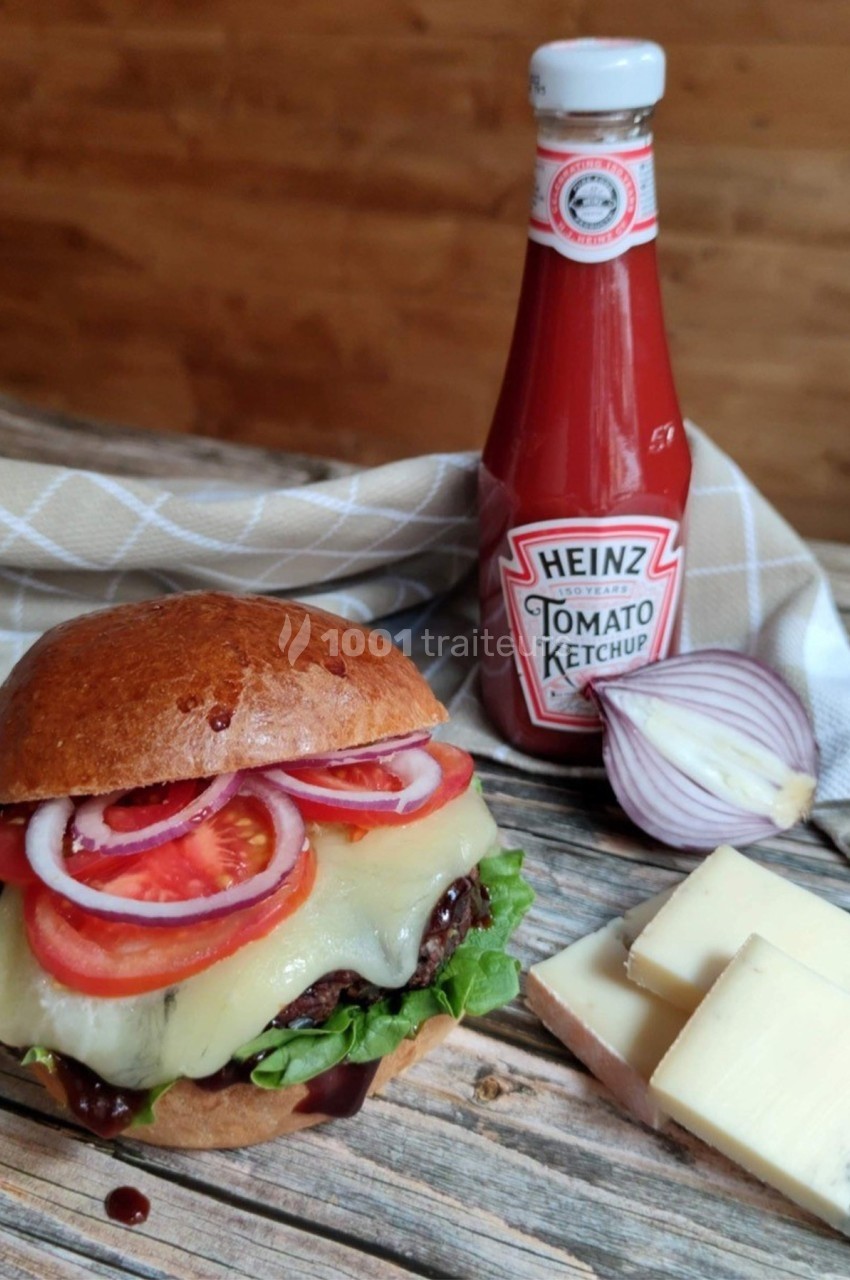 Burger garni de fromage fondu, salade, tomates et oignons rouges, avec une bouteille de ketchup et du fromage sur une table…