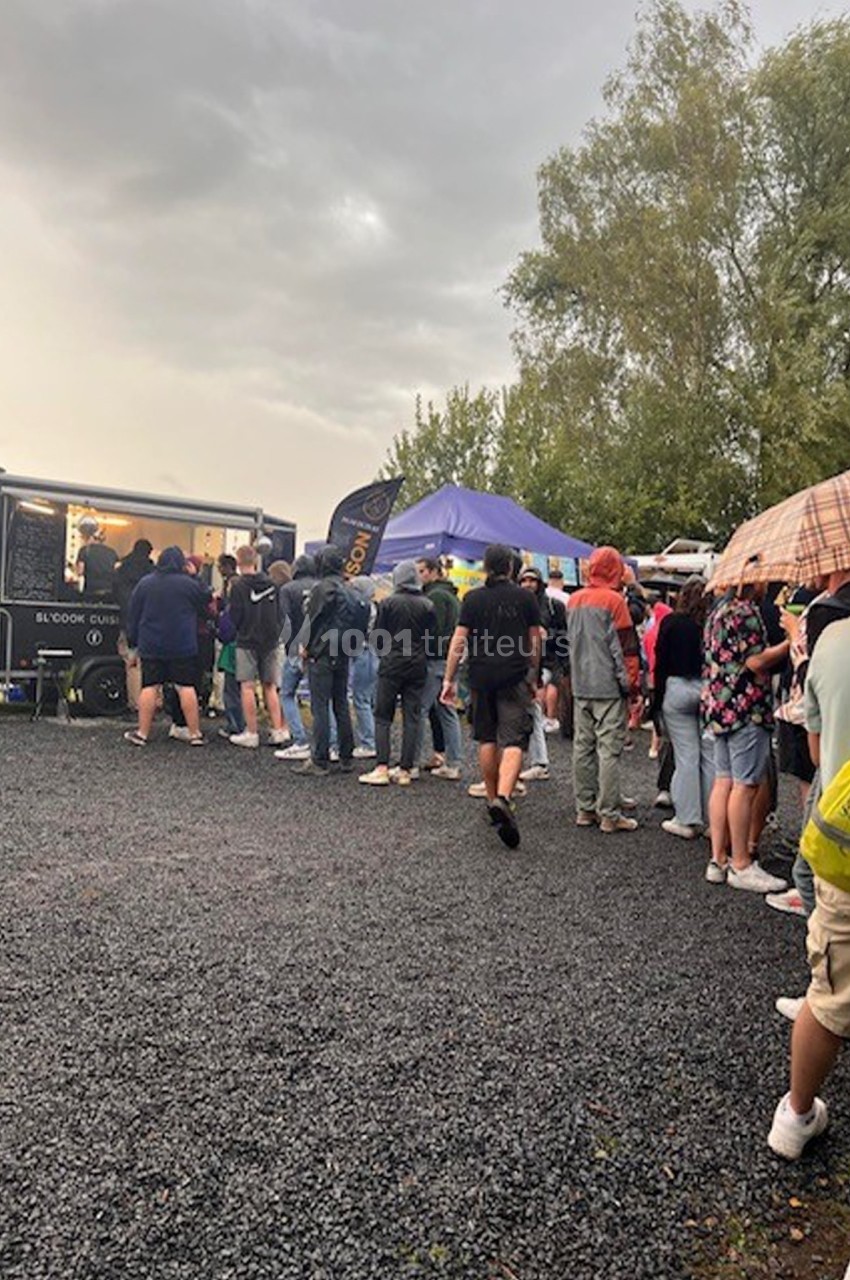 File d'attente devant des stands de nourriture en plein air par temps nuageux, avec des arbres en arrière-plan.