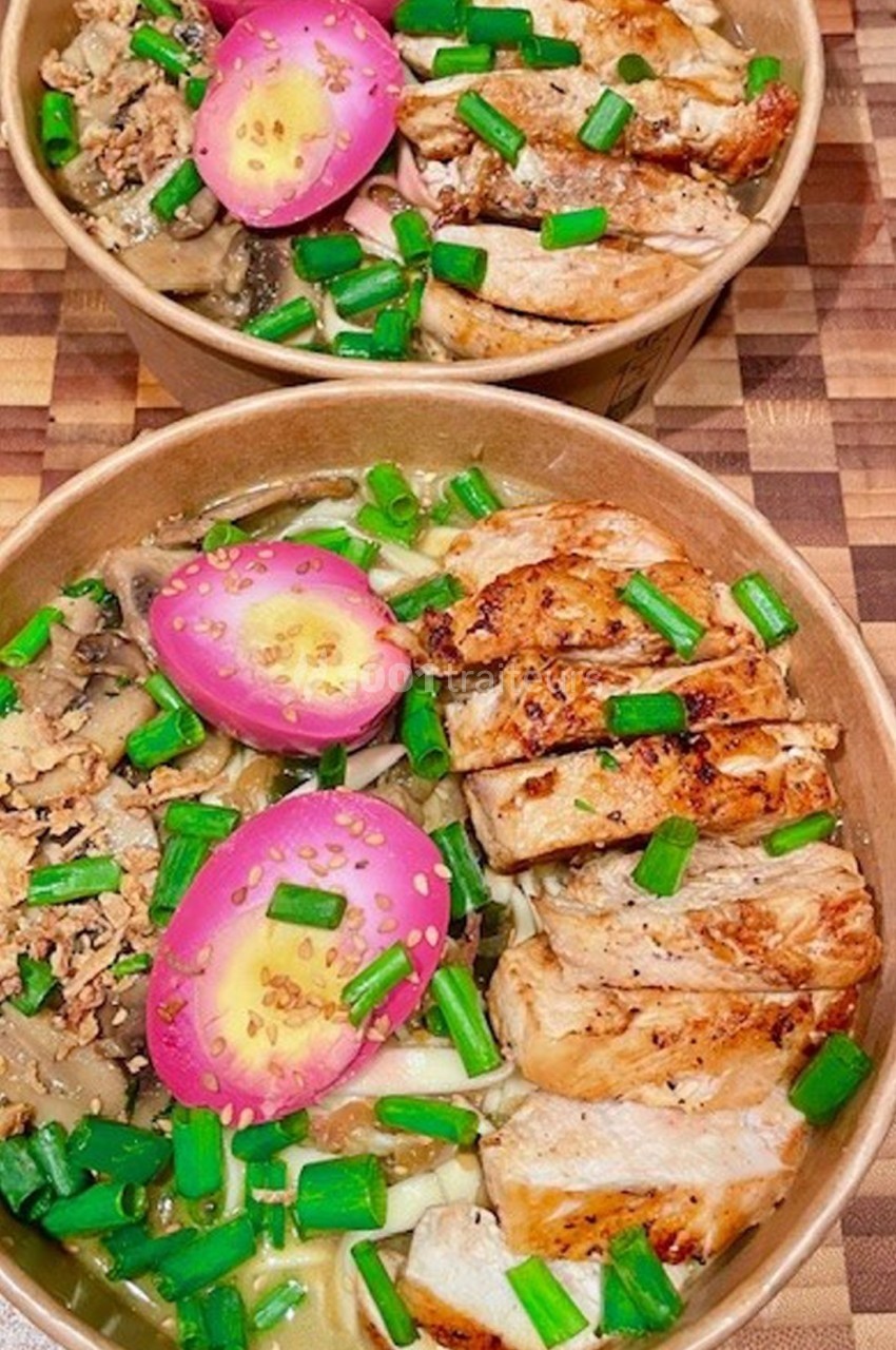 Bol de nouilles garni de poulet grillé, œufs marinés roses, champignons et oignons verts sur une table en bois.