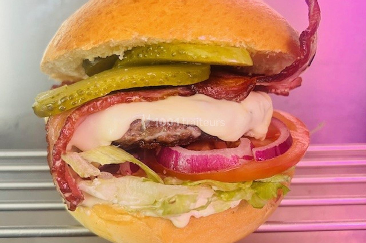 Burger garni de bacon, fromage fondu, cornichons, rondelles d'oignon, tomate et laitue, posé sur une grille métallique.