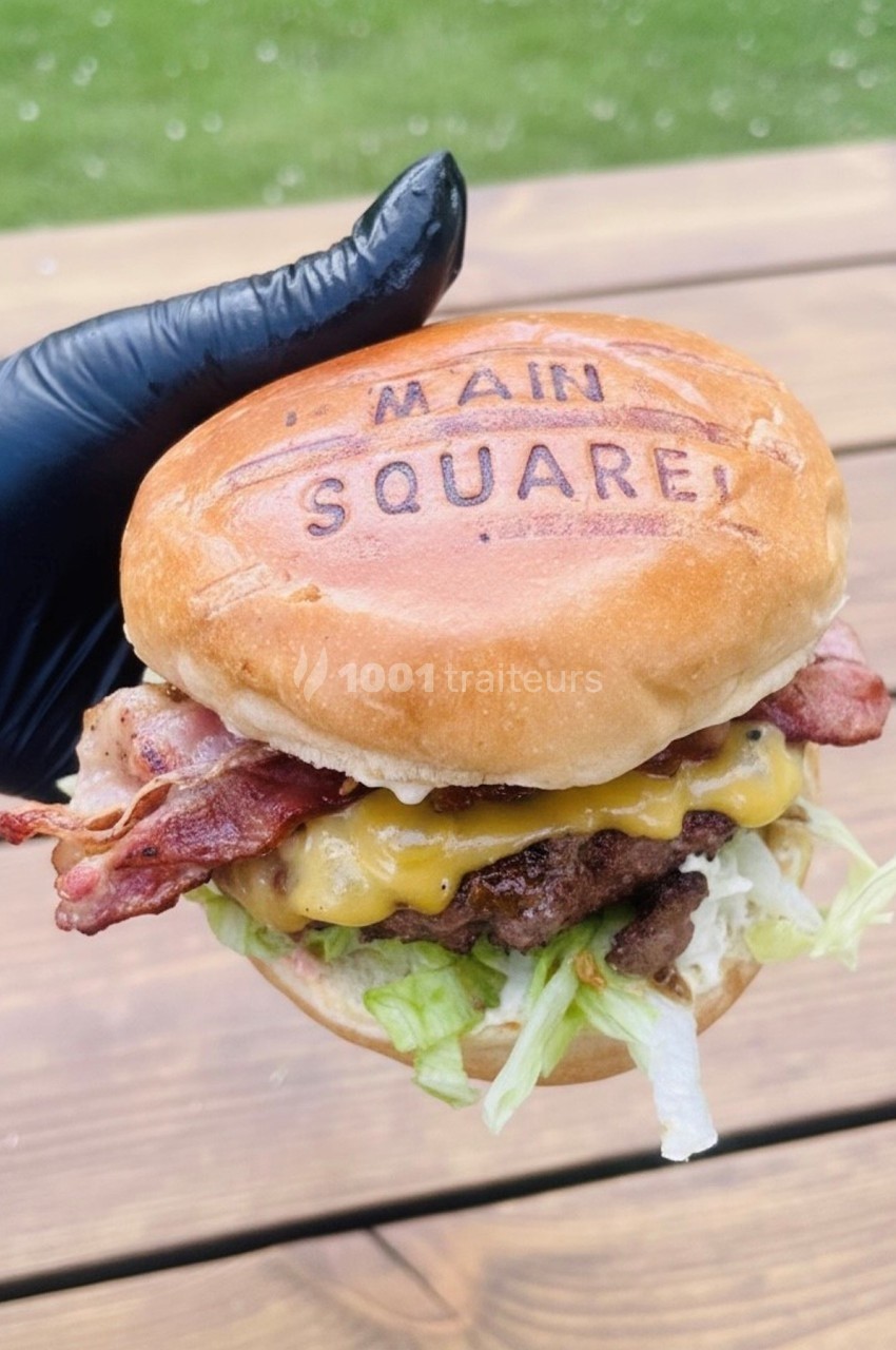 Burger garni de bacon, fromage fondu, laitue et steak, tenu par une main gantée, sur fond de table en bois.
