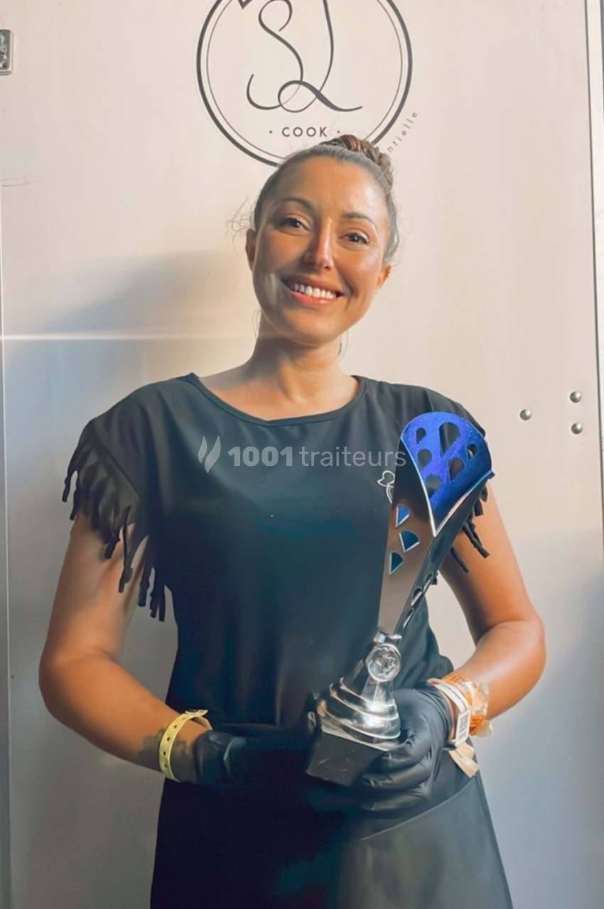 Femme souriante tenant un trophée, vêtue de noir, devant un mur clair avec un logo circulaire en arrière-plan.