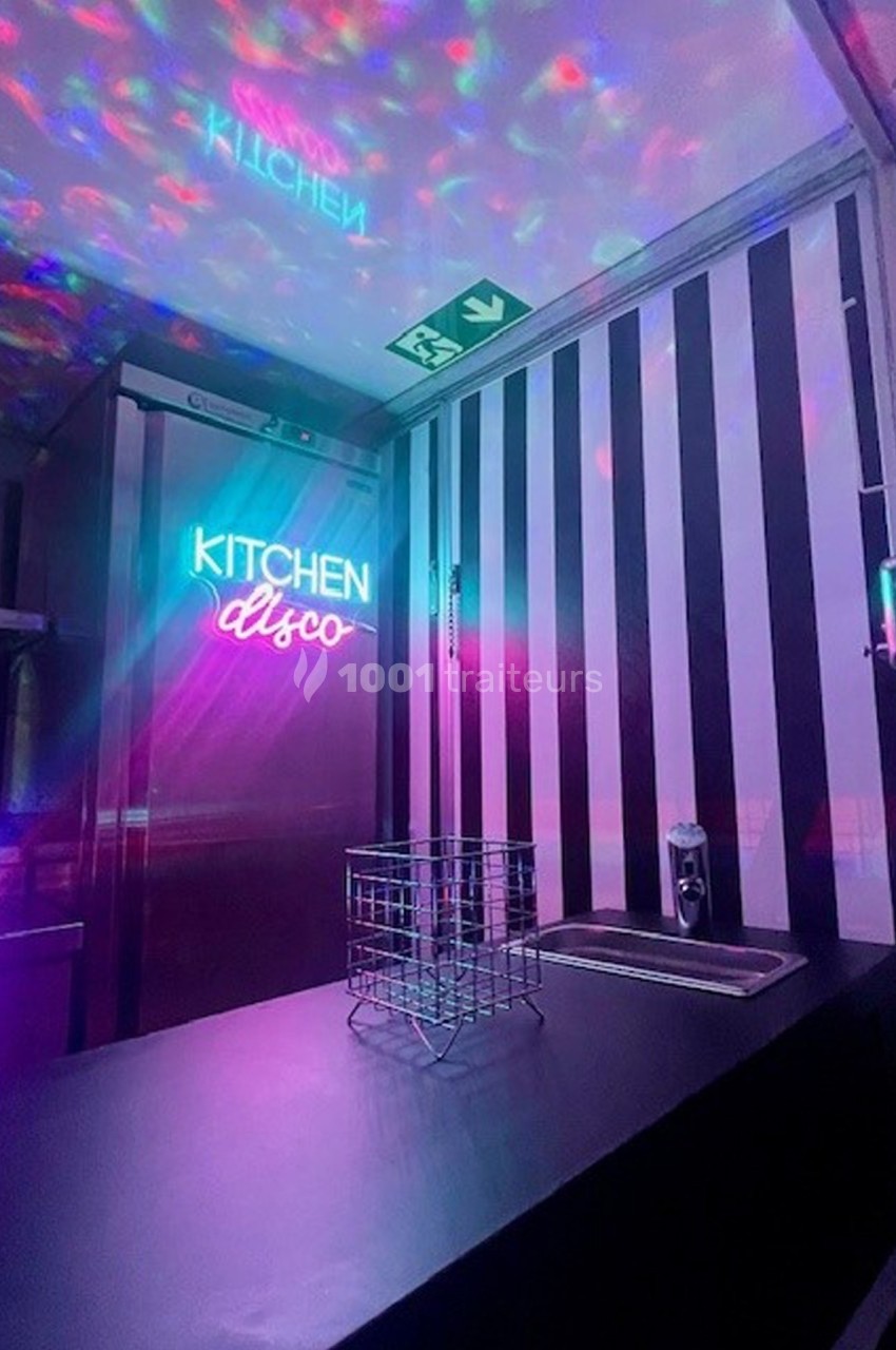 Plan de travail sombre avec un évier, décoré de lumières colorées et d'un néon ’Kitchen disco’ sur un mur rayé.