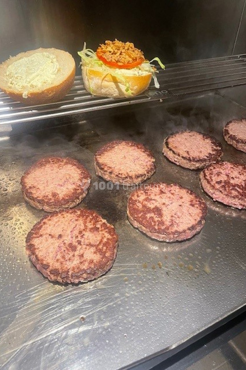 Burgers en cuisson sur une plaque chauffante avec un pain garni de légumes et sauce en arrière-plan.