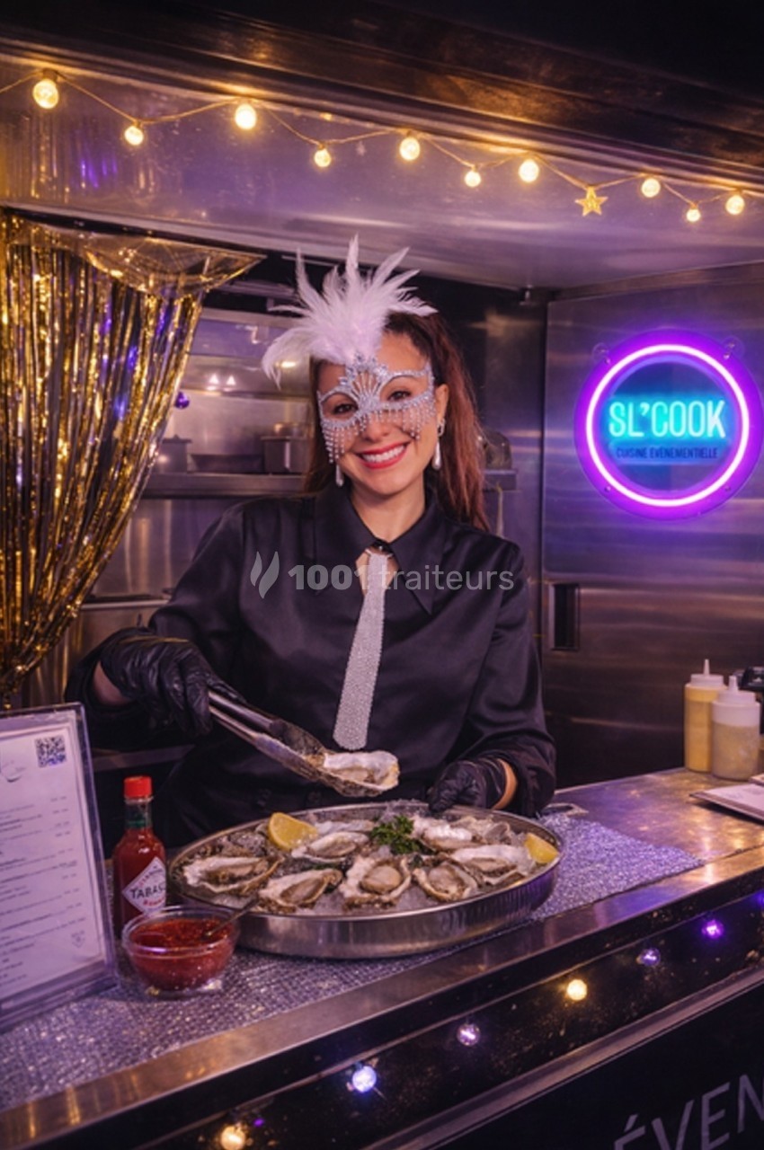 Femme masquée servant des huîtres sur un stand décoré avec des lumières et un panneau lumineux en arrière-plan.