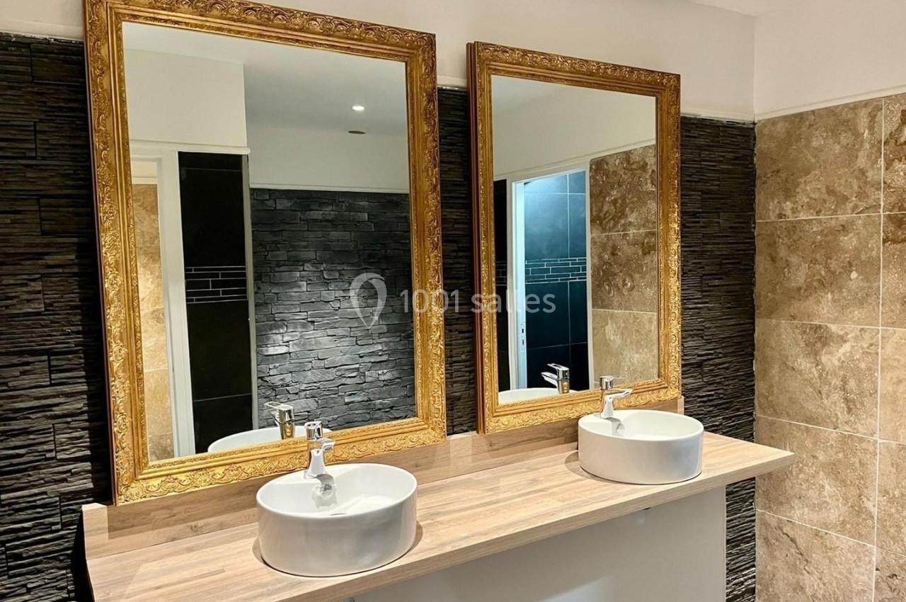 Deux lavabos blancs sur un plan en bois clair, surmontés de miroirs dorés, avec un mur en pierre et carrelage beige.