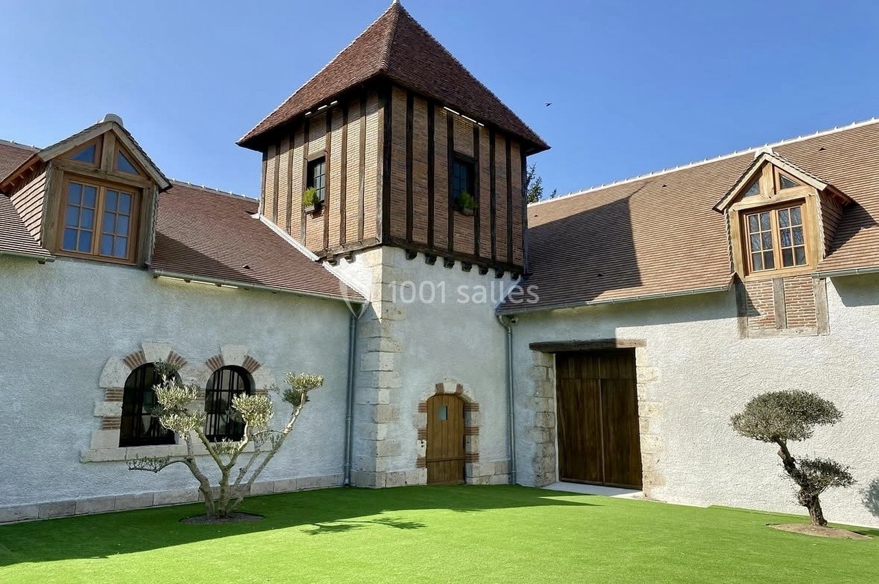 Bâtisse ancienne avec tour centrale, murs en pierre et toit en tuiles, entourée d'un jardin avec pelouse artificielle.