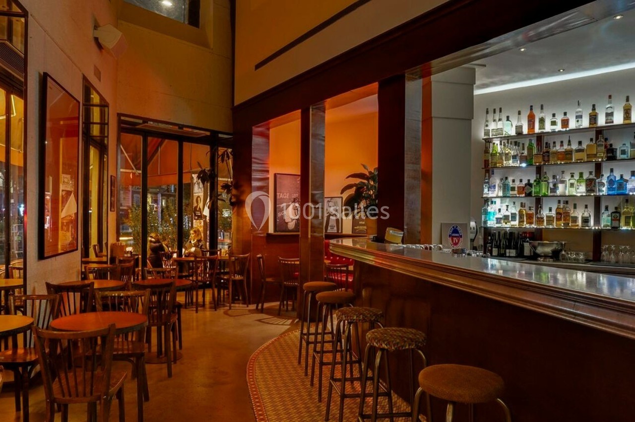 Intérieur d'un bar avec un comptoir éclairé, des étagères de bouteilles et des tables en bois dans une ambiance chaleureuse.