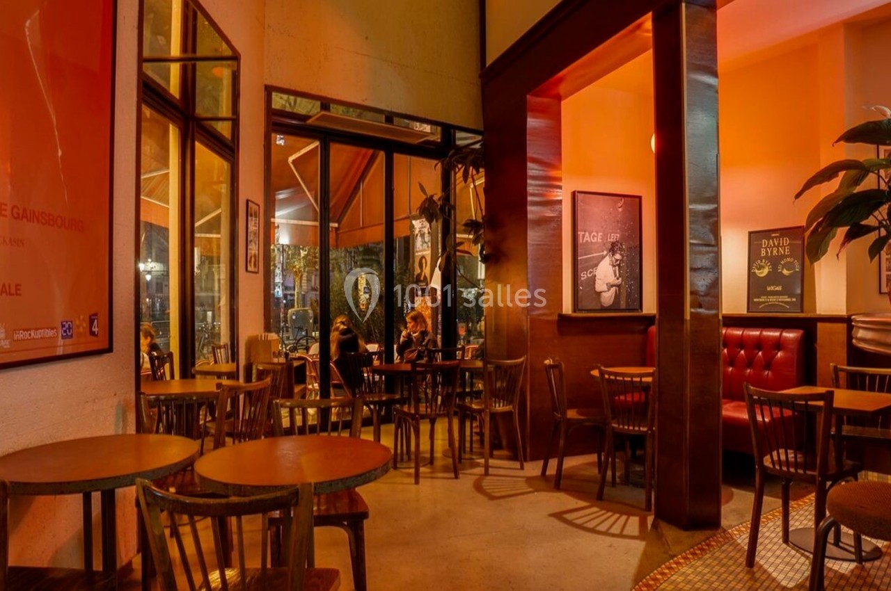 Salle de café chaleureuse avec éclairage tamisé, tables en bois, affiches aux murs et plantes en décoration.