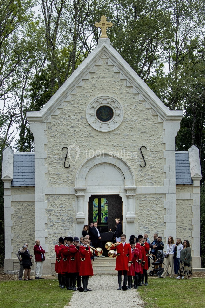 Des musiciens en uniforme rouge jouent devant une chapelle en pierre entourée de spectateurs dans un cadre boisé.