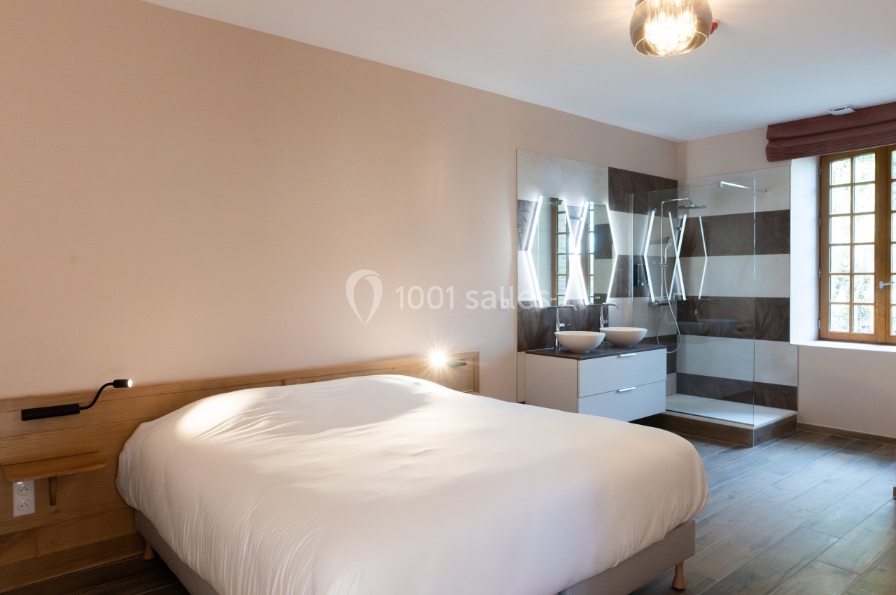 Chambre lumineuse avec lit double, salle de bain ouverte comprenant une douche vitrée et un lavabo moderne.