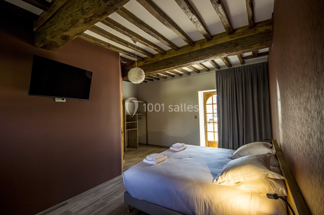 Chambre avec lit double, télévision murale, poutres apparentes et lumière naturelle entrant par une porte-fenêtre.
