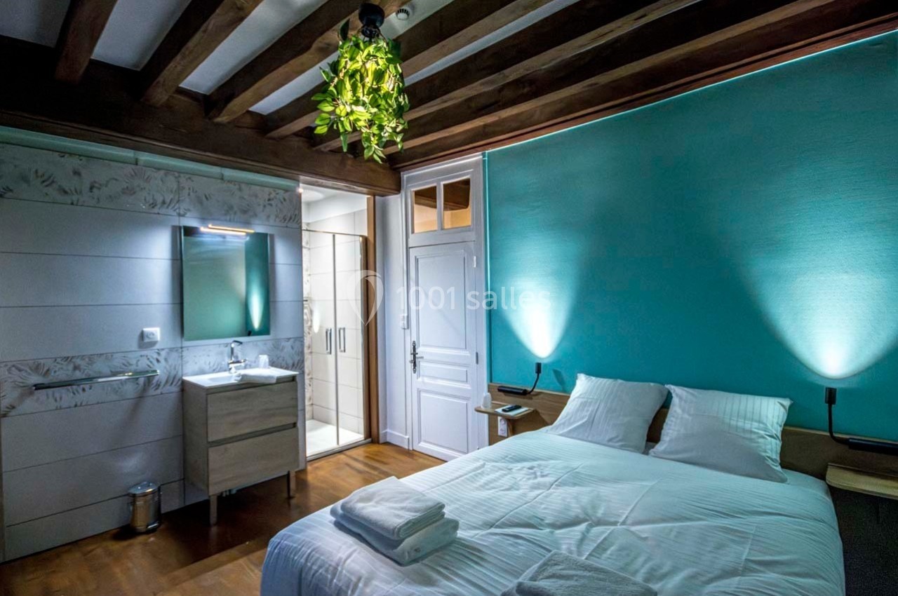 Chambre avec lit double, plafond en poutres apparentes, mur turquoise, salle de bain attenante et éclairage doux.