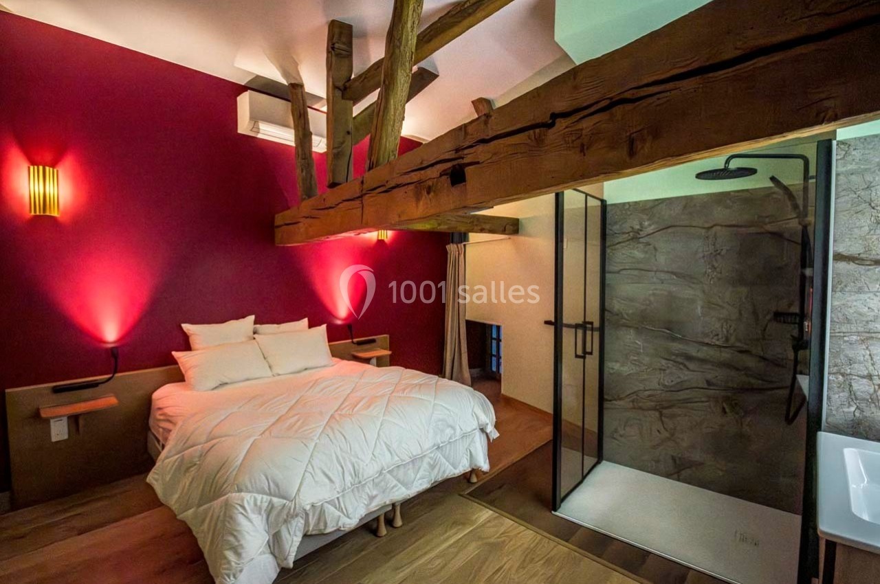 Chambre avec lit double, poutres apparentes, éclairage chaleureux et salle de douche moderne ouverte.