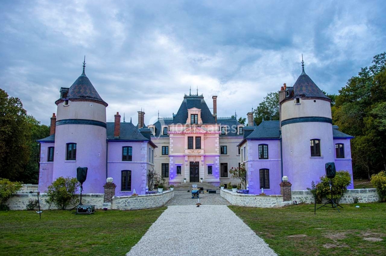 Façade d'un château éclairé de lumières violettes, entouré de tours et d'un jardin avec une allée centrale.