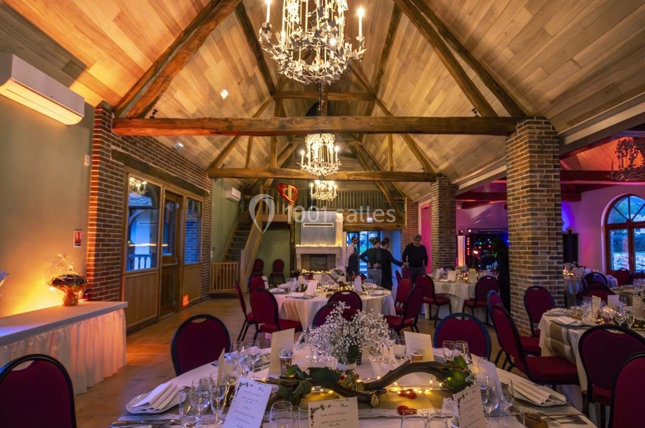 Salle de réception avec poutres apparentes, tables dressées, chaises rouges et éclairage chaleureux.
