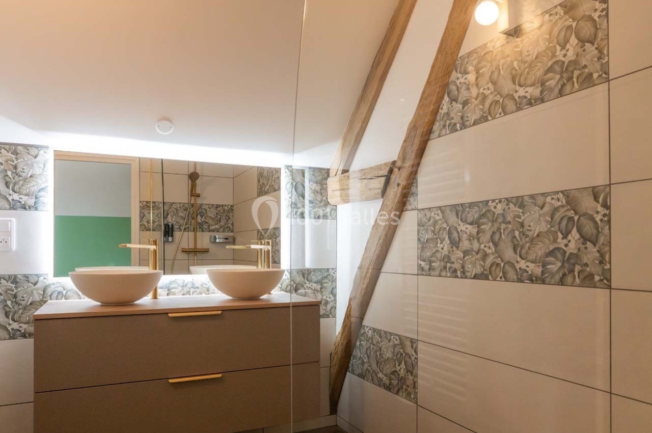Salle de bain moderne avec double vasque, miroir éclairé, carrelage floral et poutre apparente en bois.