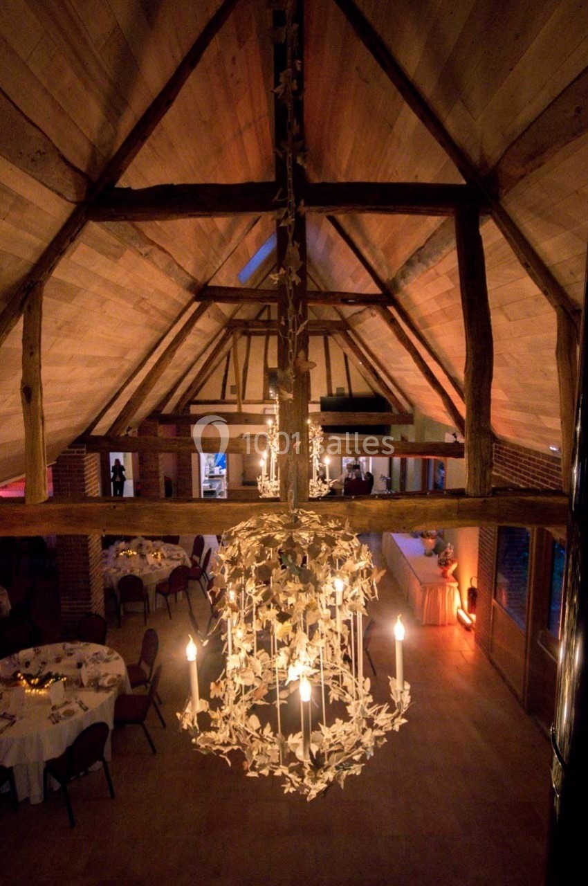 Vue intérieure d'une salle de réception avec poutres apparentes, grand lustre décoratif et tables dressées.