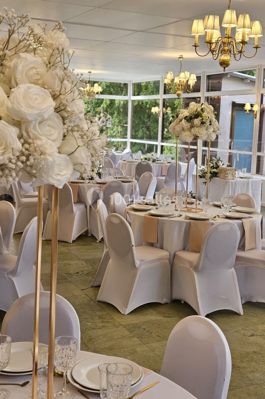 Salle de réception décorée avec des tables rondes, nappes blanches, centres de table fleuris et lustres suspendus.