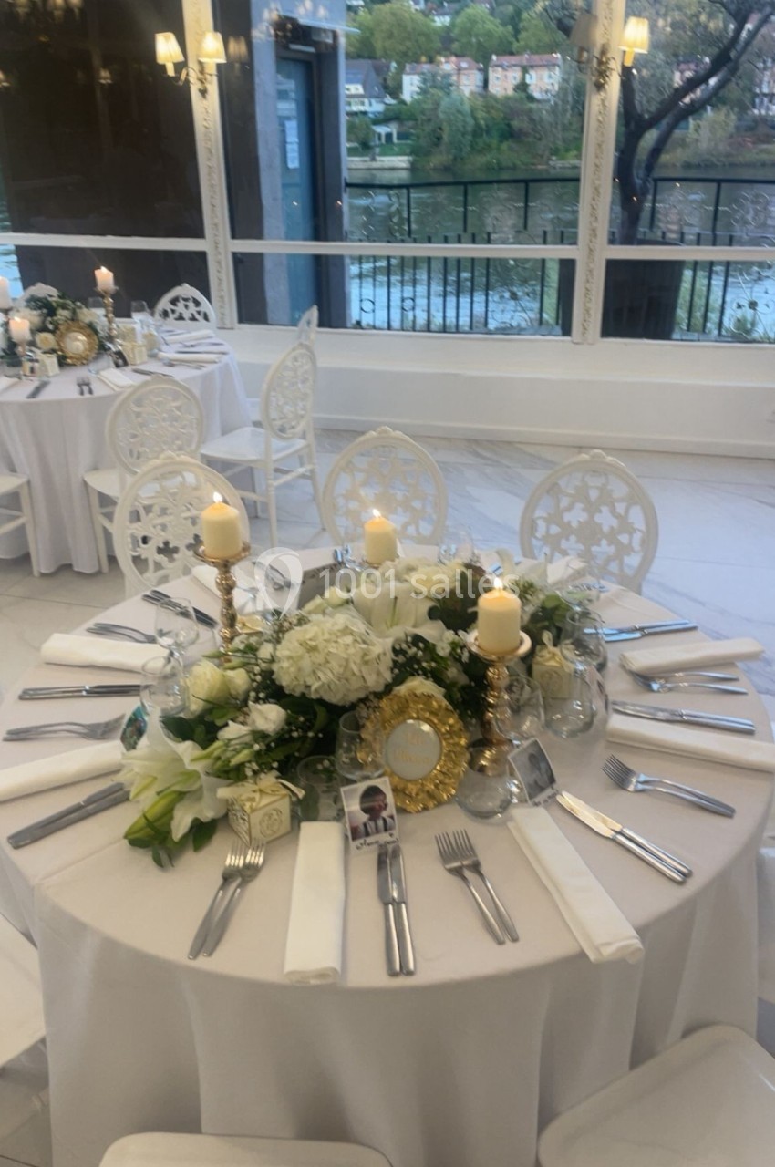 Table élégamment dressée avec des fleurs blanches, bougies et couverts, dans une salle lumineuse avec vue sur l'extérieur.