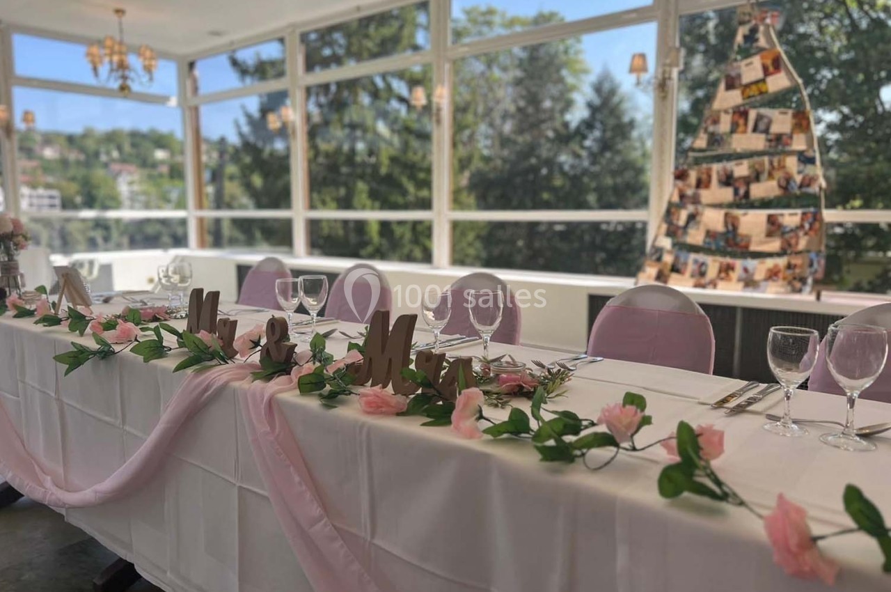 Table décorée pour un événement avec nappes blanches, fleurs roses et guirlande, dans une salle lumineuse avec vue sur des…