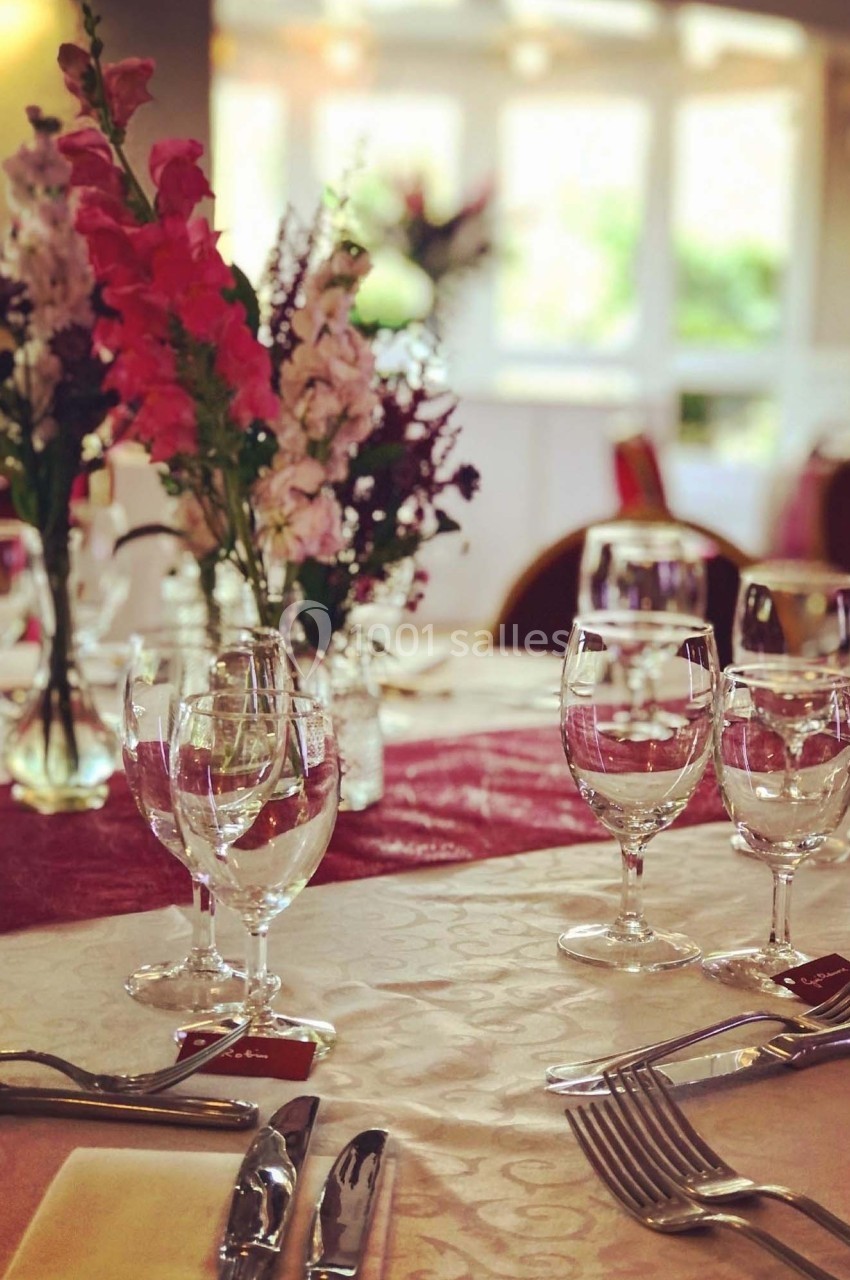 Table élégamment dressée avec nappes blanches, verres, couverts et bouquets de fleurs colorées en arrière-plan lumineux.