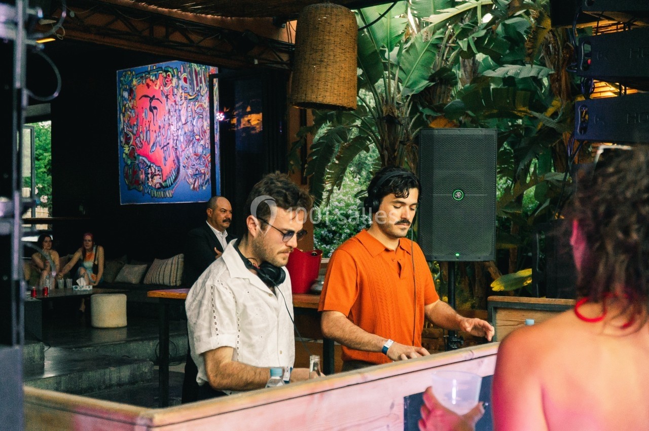 Deux DJ mixent de la musique dans un espace extérieur décoré de plantes et d'une œuvre d'art colorée au mur.
