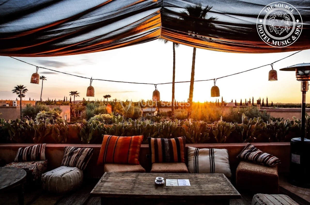 Terrasse aménagée avec des coussins et des lanternes suspendues, offrant une vue sur un coucher de soleil et des palmiers.