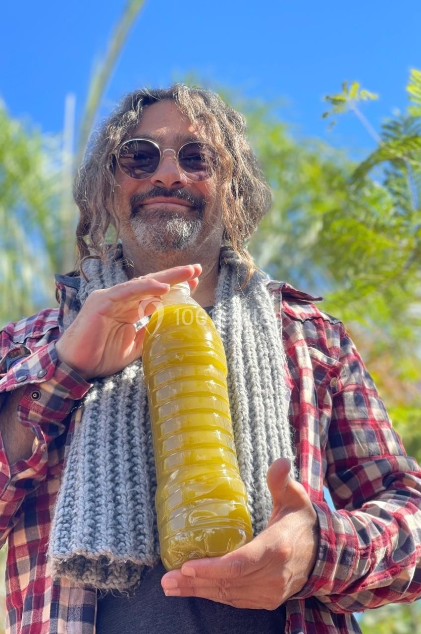 Homme souriant portant des lunettes rondes, tenant une bouteille remplie d'un liquide jaune, en extérieur sous un ciel bleu.