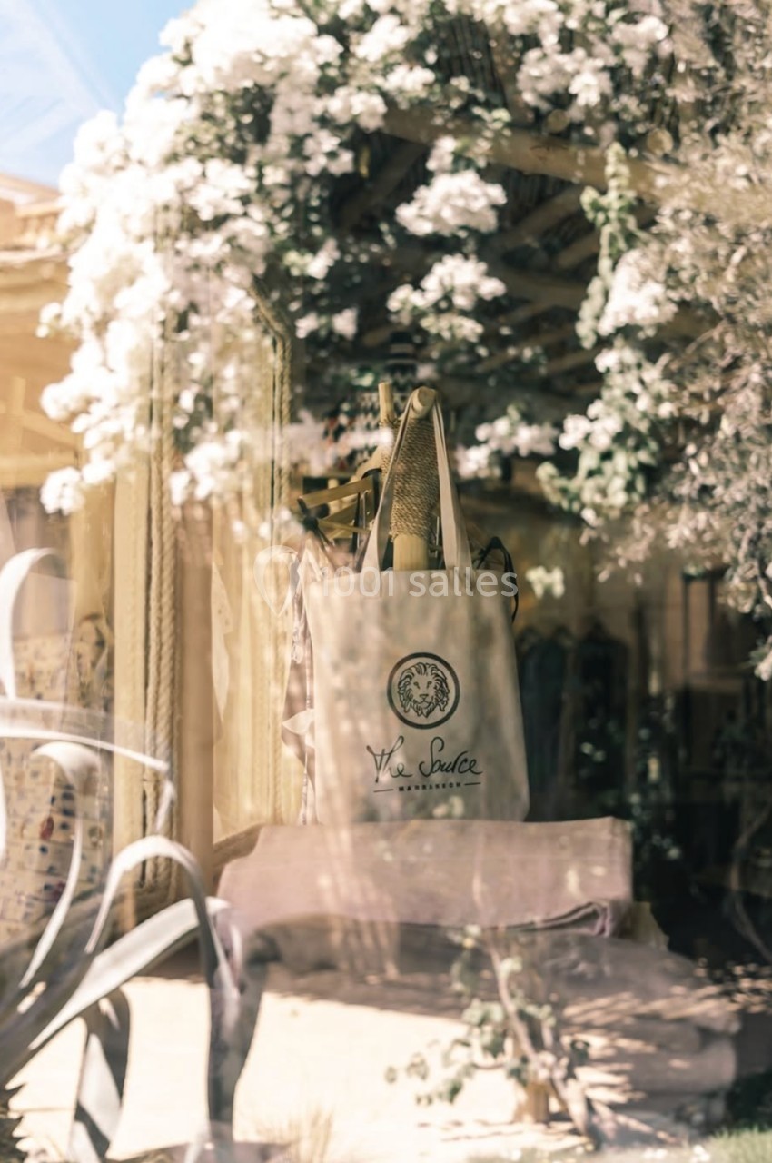 Sac en toile beige suspendu devant une vitrine, entouré de fleurs blanches et de plantes en arrière-plan.