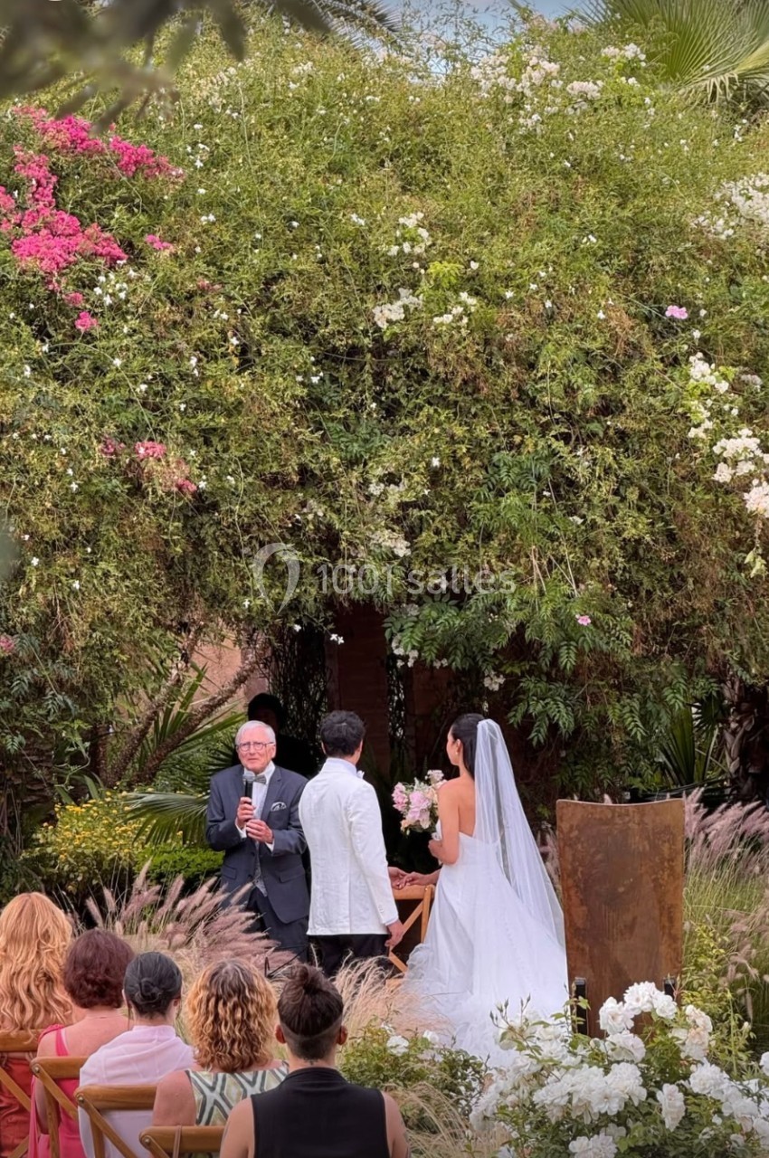 Un couple se marie en extérieur devant un officiant, entouré de végétation luxuriante et d'invités assis.
