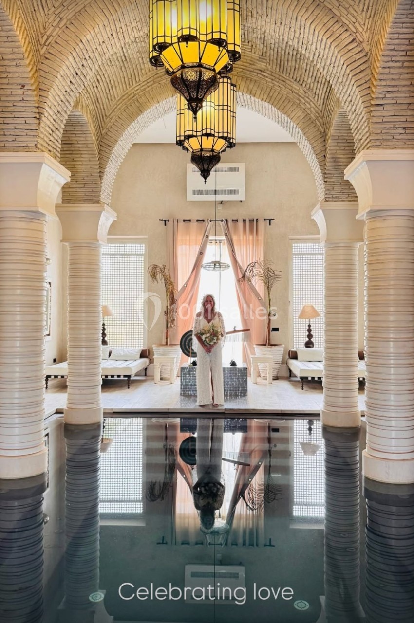 Femme en robe blanche tenant un bouquet, debout dans une salle voûtée avec piscine intérieure et lumière naturelle.