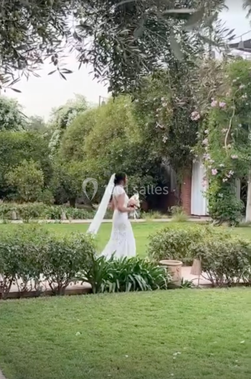 Une mariée en robe blanche et voile traverse un jardin verdoyant, tenant un bouquet de fleurs.