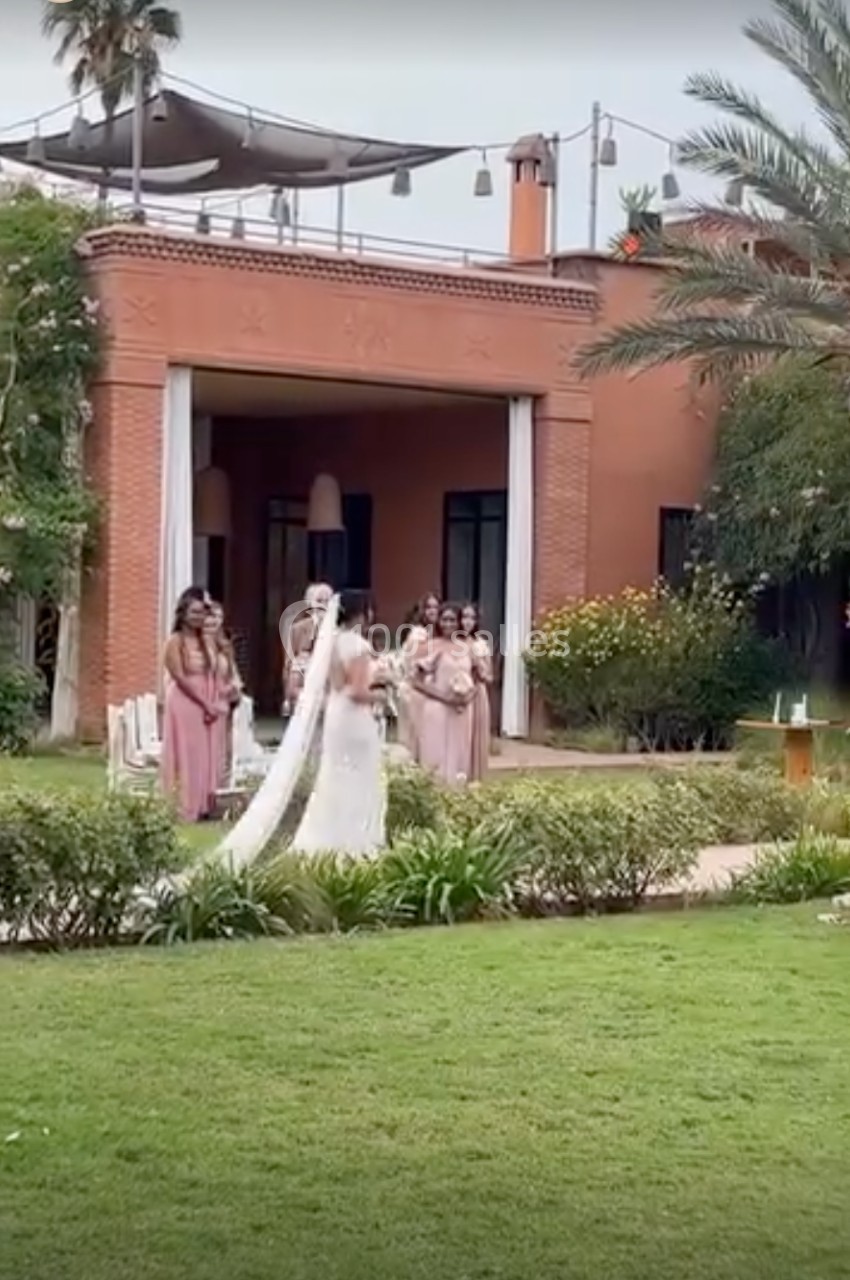 Une mariée en robe blanche marche dans un jardin verdoyant, suivie de demoiselles d'honneur en robes roses.