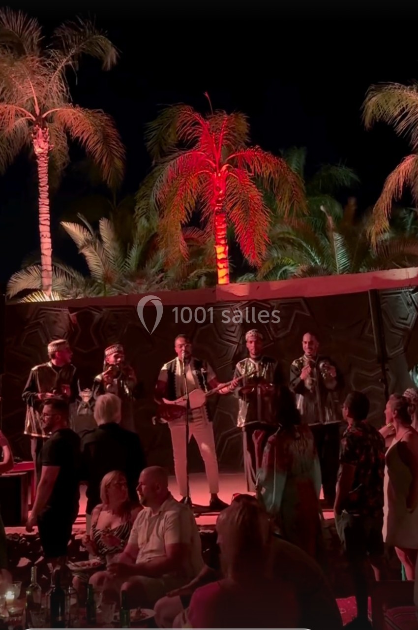 Un groupe de musiciens chante sur scène en plein air, entouré de palmiers éclairés et d'un public debout.