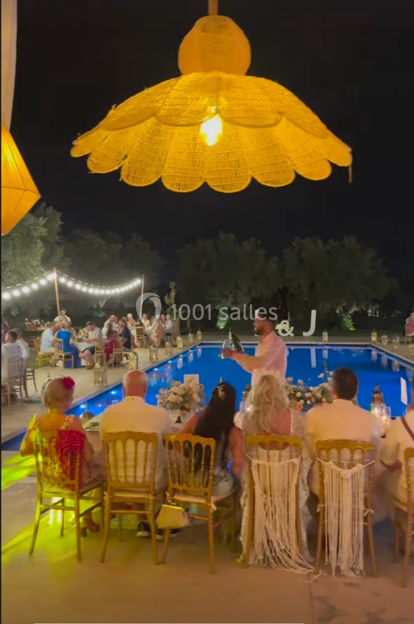 Des convives dînent en extérieur autour d'une piscine éclairée, sous des lampes suspendues, en soirée.