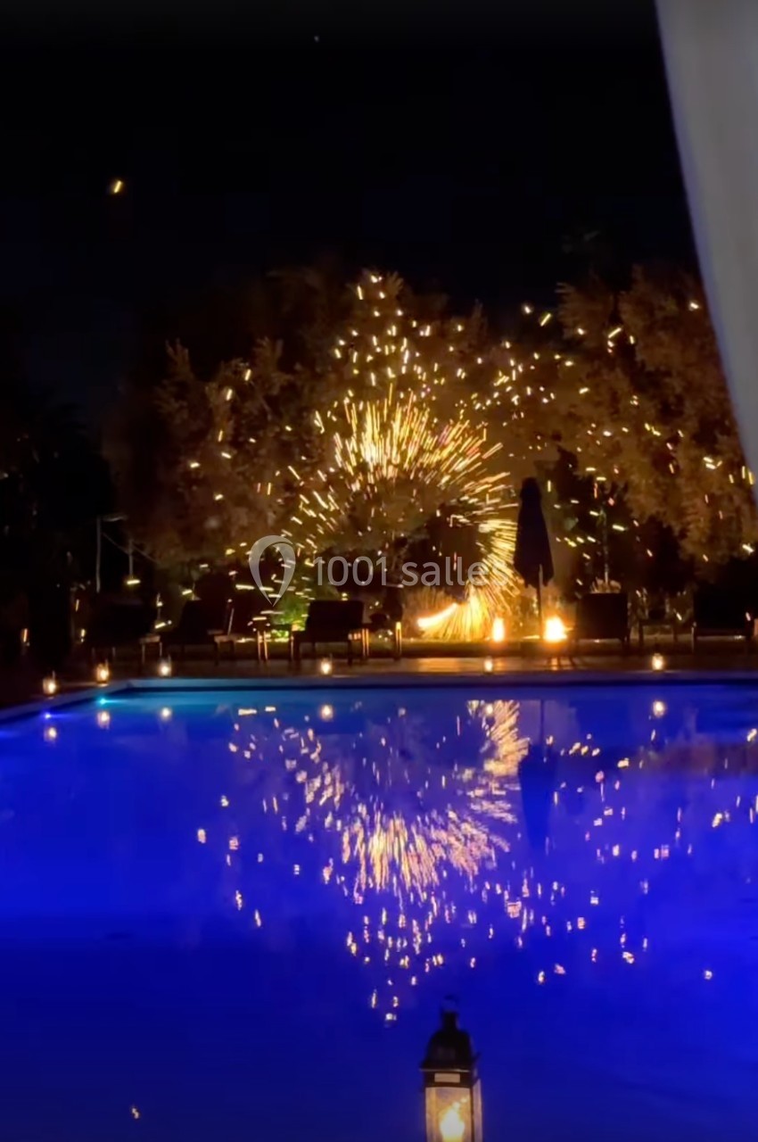 Feux d'artifice illuminant des arbres près d'une piscine éclairée de nuit, avec des reflets dans l'eau calme.