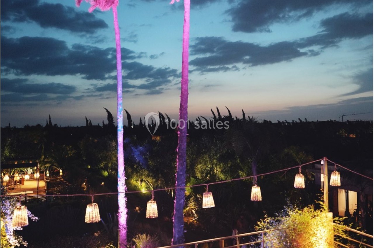 Terrasse en soirée avec des palmiers éclairés en rose, des guirlandes lumineuses et des personnes assises autour de tables.