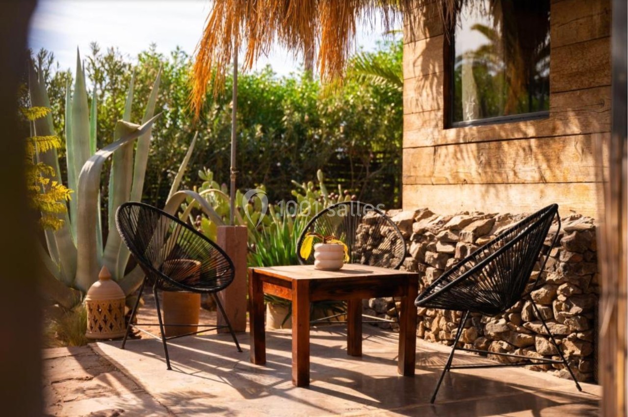 Terrasse ensoleillée avec deux fauteuils noirs, une table en bois et des plantes exotiques en arrière-plan.