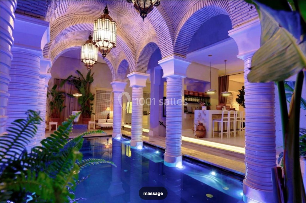 Piscine intérieure éclairée entourée de colonnes et d'arches, avec un décor exotique et un bar en arrière-plan.
