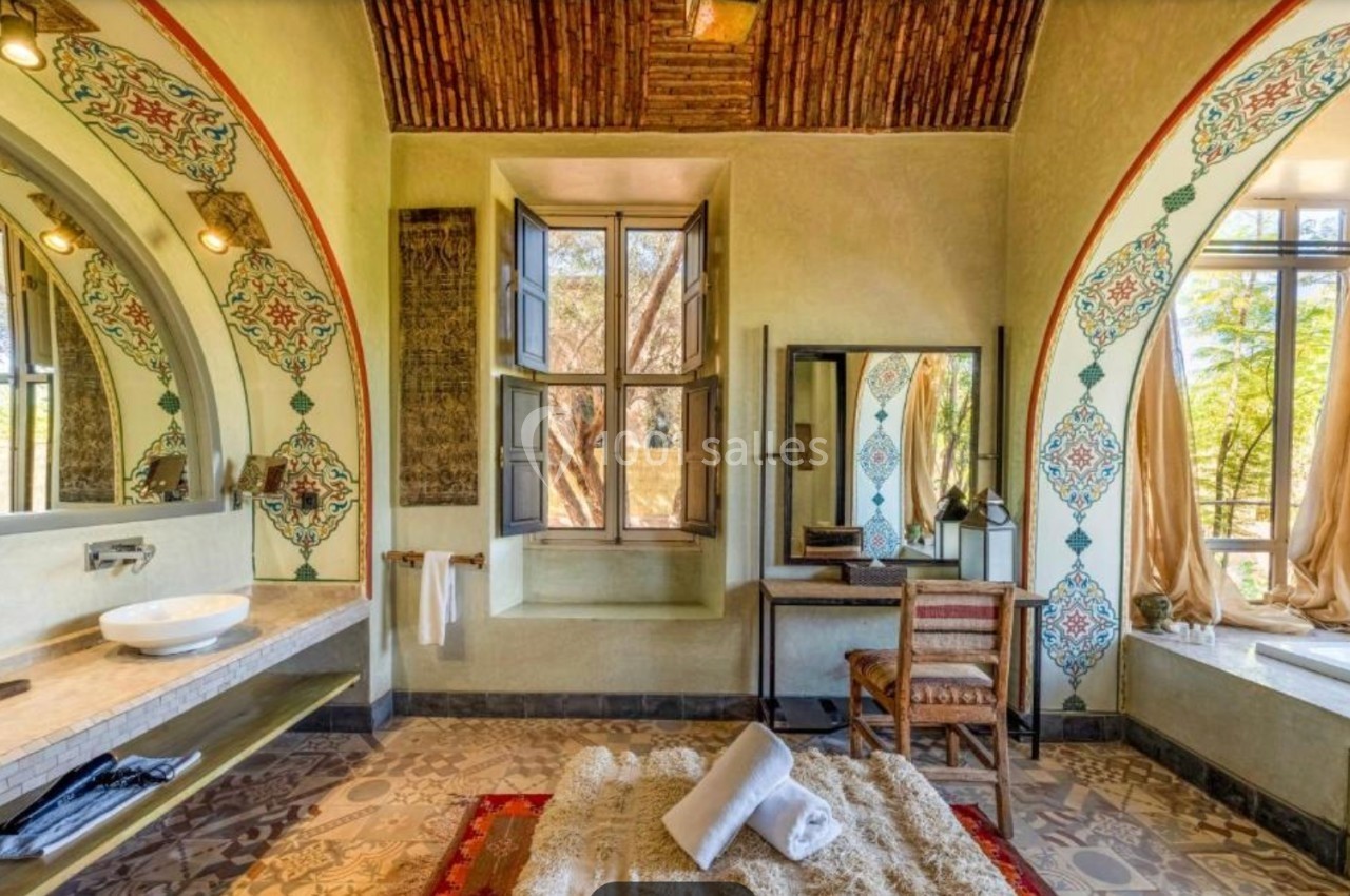 Salle de bain lumineuse avec décorations orientales, grande fenêtre, lavabo, miroir, chaise et tapis au sol.