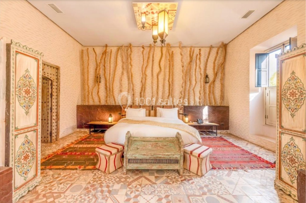 Chambre lumineuse avec lit double, tapis colorés, mobilier en bois sculpté et murs ornés de motifs naturels.