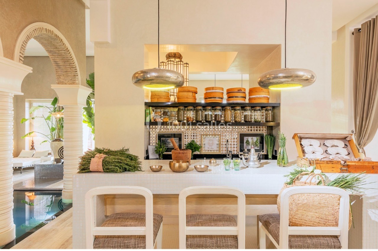 Cuisine lumineuse avec comptoir blanc, tabourets en rotin, étagères garnies de bocaux et paniers, décorée de plantes vertes.
