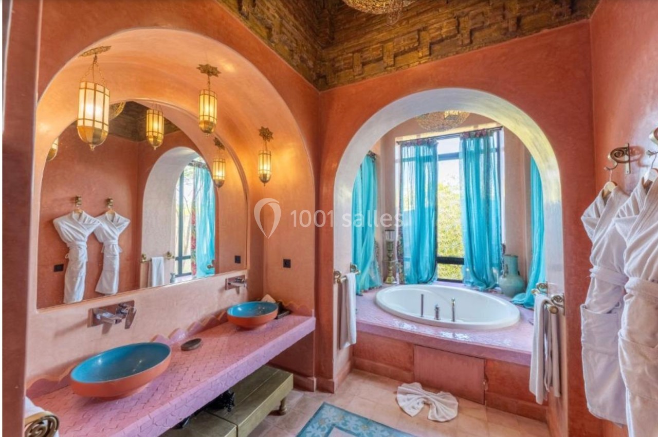 Salle de bain colorée avec murs roses, vasques bleues, baignoire encastrée, rideaux turquoise et éclairage suspendu.